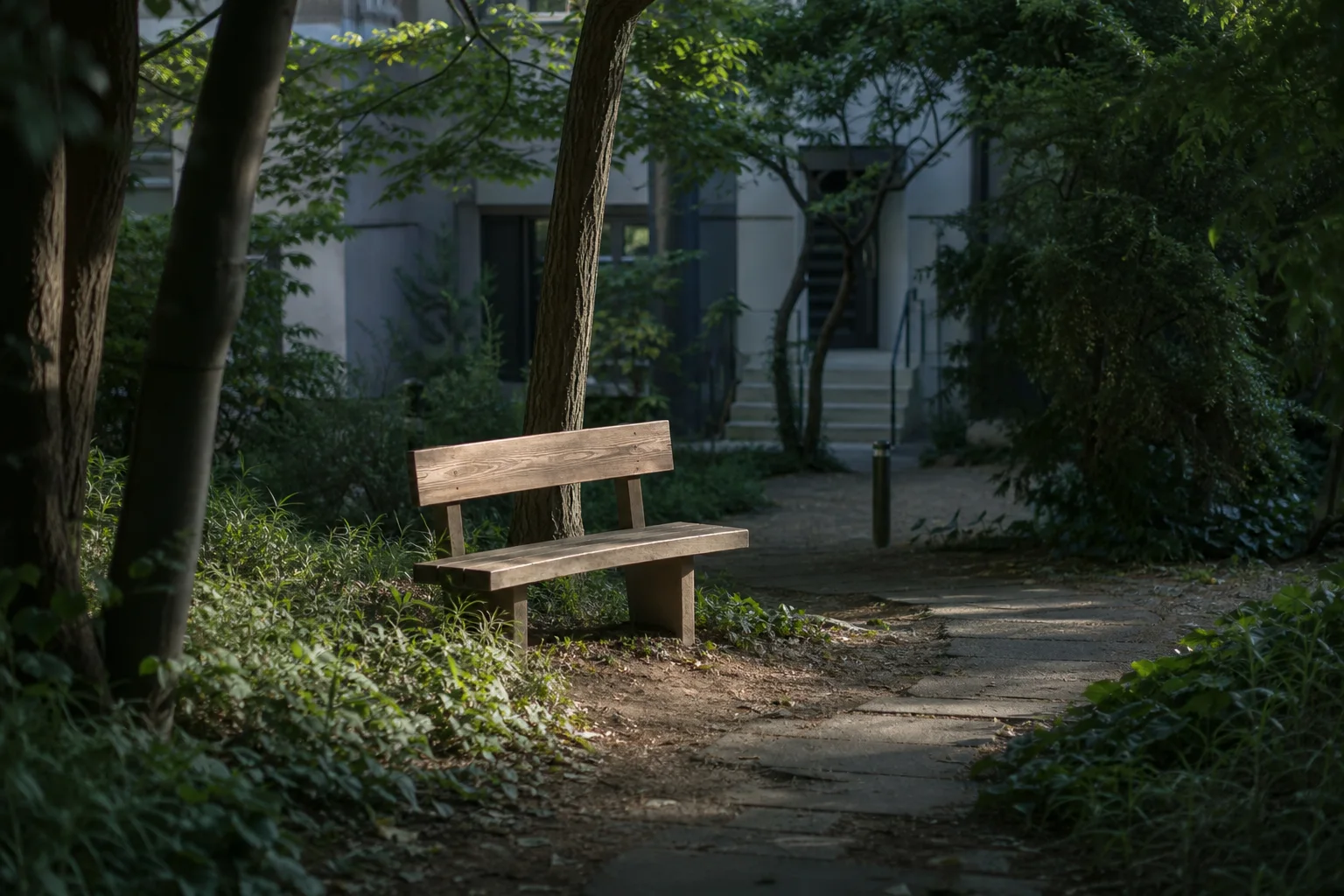 나무 그늘 아래 작은 목재 벤치가 놓이고 좁은 보행로가 주거동 입구 방향으로 이어지는 쉼터