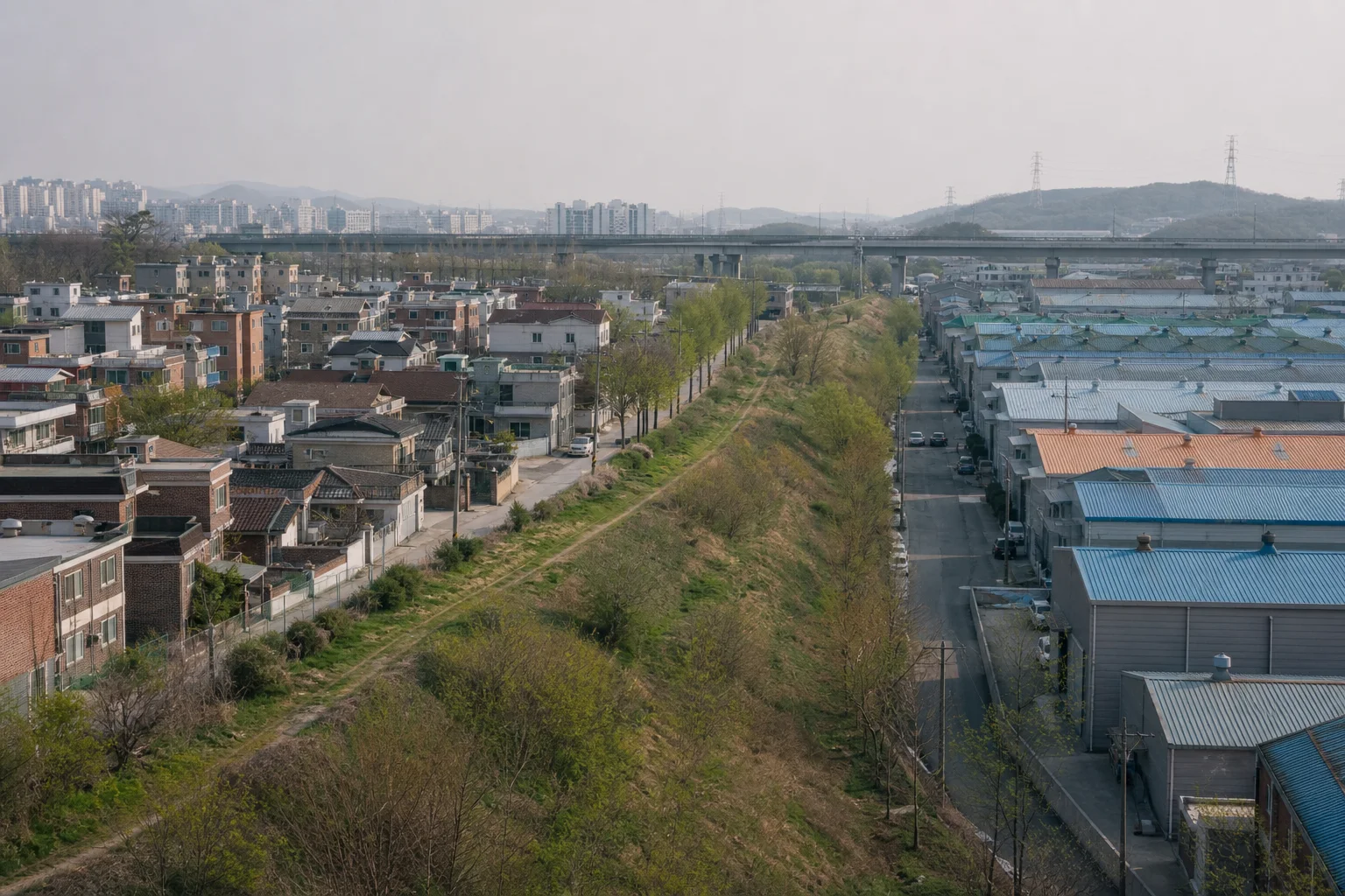 저층 주거지와 작은 공장 지붕 사이에 나무가 이어진 둑이 놓이고 멀리 고가도로와 아파트가 보이는 도시 가장자리