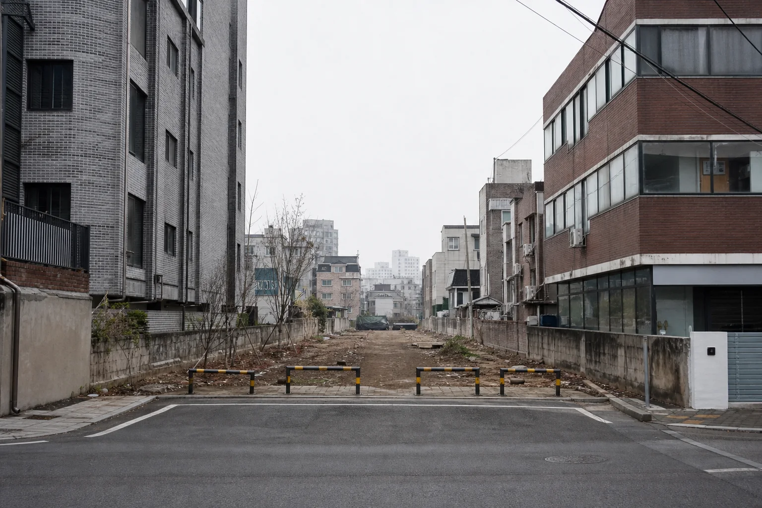 저층 건물 사이로 길게 비어 있는 흙길 형태의 공간이 직선으로 이어지고 앞쪽 도로에는 차량 차단봉이 놓인 모습